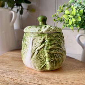 Può includere: Un barattolo in ceramica a forma di testa di cavolo, con coperchio abbinato e un piccolo pomello verde. Il barattolo è principalmente verde con accenti bianchi e poggia su una superficie di legno. Articoli da cucina decorativi.