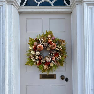 May include: A circular autumn wreath featuring a collection of small pumpkins in shades of orange, cream, and muted brown, accented with green and rust-colored foliage, white berries, and dried leaves, mounted on a light gray paneled front door.