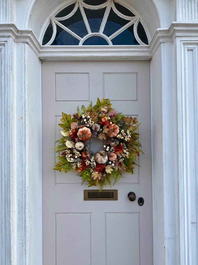 May include: A circular autumn wreath featuring a collection of small pumpkins in shades of orange, cream, and muted brown, accented with green and rust-colored foliage, white berries, and dried leaves, mounted on a light gray paneled front door.