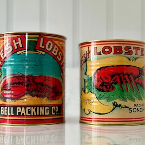 May include: Two vintage tin cans of fresh lobster. The cans feature colorful labels with a red lobster illustration, the words "FRESH LOBSTER", and "BELL PACKING CO." The cans are cylindrical and have a retro design.