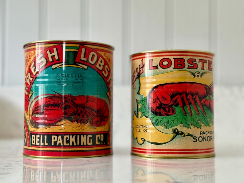 May include: Two vintage tin cans of fresh lobster. The cans feature colorful labels with a red lobster illustration, the words "FRESH LOBSTER", and "BELL PACKING CO." The cans are cylindrical and have a retro design.