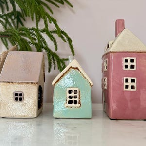 Peut inclure: Trois figurines de maisons en c&eacute;ramique aux couleurs pastel. Les maisons ont des fen&ecirc;tres carr&eacute;es et des toits de formes diff&eacute;rentes. Une est rose clair, une est bleu clair et une est blanc cass&eacute; avec un toit marron clair. D&eacute;coratives, elles conviennent &agrave; la d&eacute;coration int&eacute;rieure.