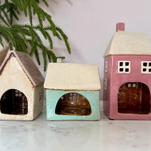 Peut inclure: Trois bougeoirs en c&eacute;ramique en forme de maison, cr&egrave;me, turquoise et rose. Chacun a un toit contrastant et une ouverture arqu&eacute;e. La maison rose a deux fen&ecirc;tres carr&eacute;es. D&eacute;coration int&eacute;rieure.