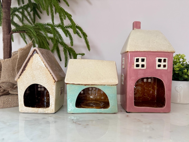 Peut inclure: Trois bougeoirs en c&eacute;ramique en forme de maison, cr&egrave;me, turquoise et rose. Chacun a un toit contrastant et une ouverture arqu&eacute;e. La maison rose a deux fen&ecirc;tres carr&eacute;es. D&eacute;coration int&eacute;rieure.