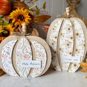Könnte beinhalten: Zwei Holz-Kürbisdekorationen mit floral gemusterten Einsätzen. Auf einem Kürbis steht "Hello Autumn" und auf dem anderen "Cosy up". Die Kürbisse sind mit Bindfäden und Holzakzenten verziert, mit Sonnenblumen im Hintergrund.