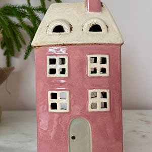 Peut inclure: Un photophore en c&eacute;ramique rose en forme de maison avec un toit et une chemin&eacute;e blancs. La maison a quatre fen&ecirc;tres carr&eacute;es et une porte grise. Objet d&eacute;coratif pour la maison.