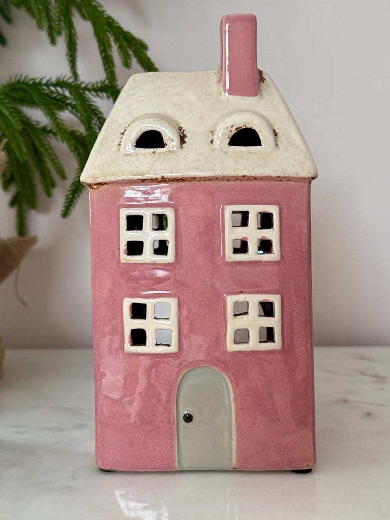 Peut inclure: Un photophore en c&eacute;ramique rose en forme de maison avec un toit et une chemin&eacute;e blancs. La maison a quatre fen&ecirc;tres carr&eacute;es et une porte grise. Objet d&eacute;coratif pour la maison.