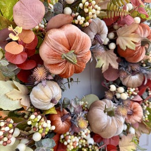May include: A decorative wreath featuring velvet pumpkins in shades of peach, tan, and rust, accented with artificial autumn leaves, sprigs of eucalyptus, and clusters of small white and cream berries arranged in a circular floral display.