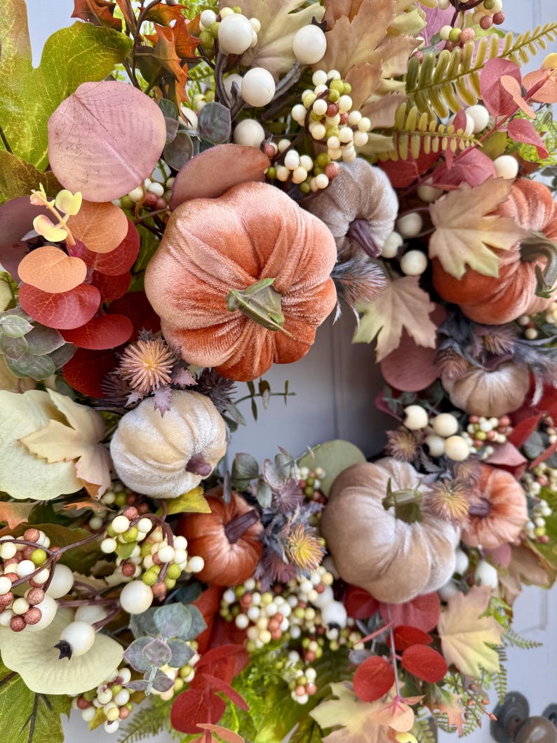May include: A decorative wreath featuring velvet pumpkins in shades of peach, tan, and rust, accented with artificial autumn leaves, sprigs of eucalyptus, and clusters of small white and cream berries arranged in a circular floral display.