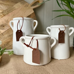May include: Three white ceramic vases with brown leather tags that say "For you". The vases are on a brown hessian surface.