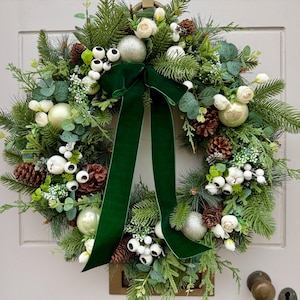 Pode incluir: Uma coroa de Natal festiva com um arranjo exuberante de galhos de sempre-viva, pinhas, bagas brancas e enfeites prateados. Uma fita de veludo verde escuro é amarrada em um laço. A coroa está em uma porta clara.