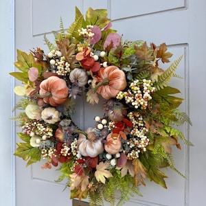 May include: A decorative wreath featuring velvet-textured pumpkins in shades of orange and cream, accented with artificial maple leaves, green ferns, eucalyptus sprigs, and clusters of small white and light green berries arranged in a circular pattern.