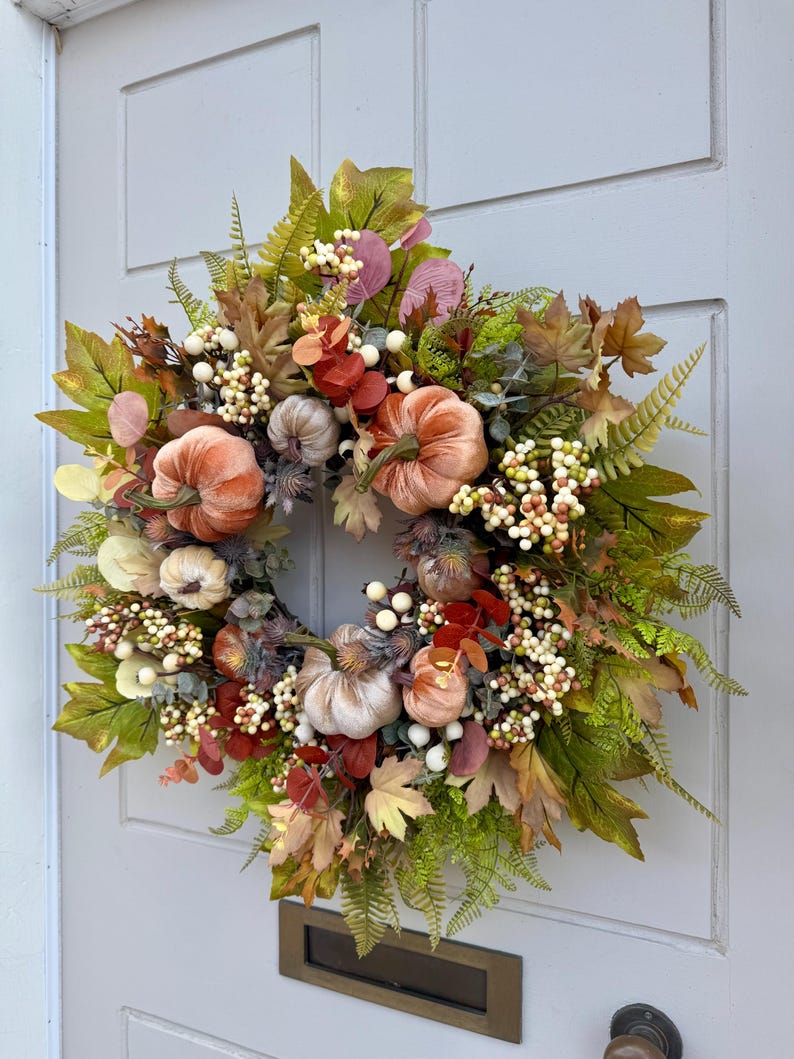 May include: A decorative wreath featuring velvet-textured pumpkins in shades of orange and cream, accented with artificial maple leaves, green ferns, eucalyptus sprigs, and clusters of small white and light green berries arranged in a circular pattern.