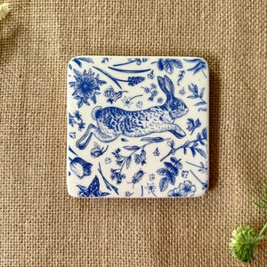 May include: A white ceramic coaster with a blue and white design of a rabbit running through a field of flowers.