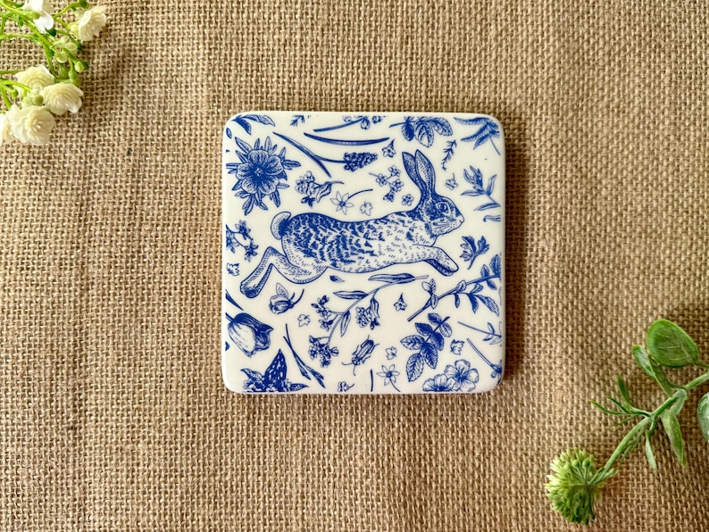 May include: A white ceramic coaster with a blue and white design of a rabbit running through a field of flowers.