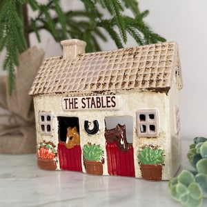 May include: A ceramic stable-shaped candle holder with the text "THE STABLES" above the stable doors. The roof is textured, and the doors have horse heads and potted plants. The color scheme is cream, brown, and red.