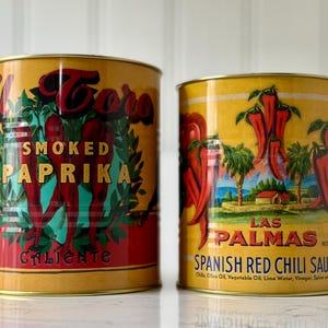 May include: Two vintage-style food cans. The left can is labeled "El Toro Smoked Paprika Caliente" with a red and green design. The right can is labeled "Las Palmas Spanish Red Chili Sauce" with a landscape scene and red chili peppers.