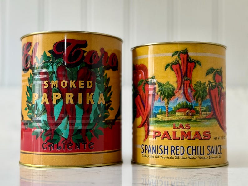 May include: Two vintage-style food cans. The left can is labeled "El Toro Smoked Paprika Caliente" with a red and green design. The right can is labeled "Las Palmas Spanish Red Chili Sauce" with a landscape scene and red chili peppers.