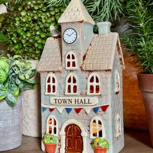 Peut inclure: Une figurine en céramique représentant un hôtel de ville avec une finition émaillée bleu clair, une tour d'horloge centrale et une porte cintrée marron. La structure présente des guirlandes rouges et bleues, des fenêtres blanches et deux pots orange.