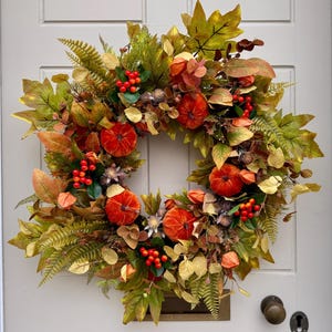 May include: A circular autumn wreath featuring a dense arrangement of faux maple leaves in shades of green, yellow, and brown, interspersed with small orange pumpkins, clusters of bright red berries, and dried fern fronds on a light gray door.