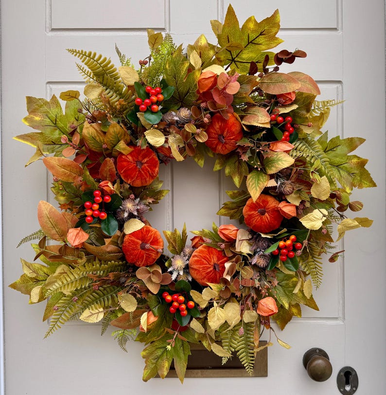 May include: A circular autumn wreath featuring a dense arrangement of faux maple leaves in shades of green, yellow, and brown, interspersed with small orange pumpkins, clusters of bright red berries, and dried fern fronds on a light gray door.
