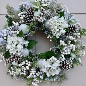 Peut inclure: Une couronne circulaire composée de fleurs d'hortensia blanches, de pommes de pin givrées et de grappes de petites baies blanches. L'arrangement comprend des feuilles vertes et des branches de sapin floquées, créant un décor hivernal dense.