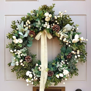 Peut inclure: Une couronne circulaire composée de feuillage vert, de baies blanches, de petites fleurs blanches et de pommes de pin brunes. Un long ruban en tissu couleur crème est suspendu au centre supérieur et tombe au milieu de la couronne sur une porte claire.
