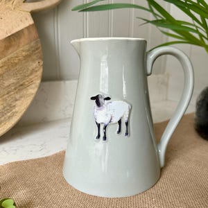 Op de afbeelding: Een lichtgrijze keramische kan met een handvat, met een schaapsticker. De kan heeft een brede basis en een smalle tuit. Het schaap is wit met een zwarte kop en poten. De kan staat op een jute oppervlak.