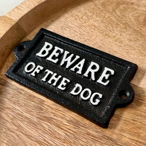Peut inclure: Panneau en fonte noire avec l'inscription "BEWARE OF THE DOG" en lettres blanches. Le panneau est de forme rectangulaire avec des coins arrondis et deux trous de fixation.