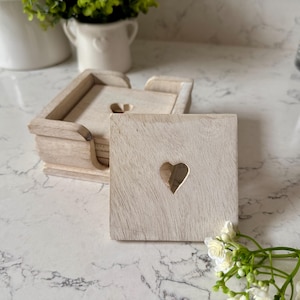 May include: Set of white wooden coasters with a heart cut-out design. The coasters are stacked in a holder and are on a white marble surface.