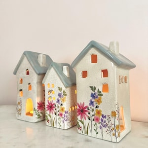 Könnte beinhalten: Drei keramikhausförmige Kerzenhalter. Die Häuser sind weiß mit blauen Dächern und mit bunten Blumenmustern verziert. Die Fenster und Türen sind ausgeschnitten, so dass das Licht durchscheinen kann. Die Kerzenhalter sind auf einer weißen Oberfläche angeordnet.