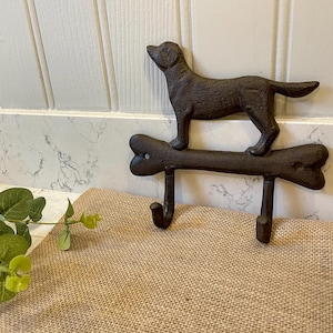 May include: A dark brown, cast metal wall hook featuring a dog silhouette standing on top of a horizontal bone shape. The bone has two downward-facing hooks attached to its underside, with visible screw holes on each end of the bone structure.