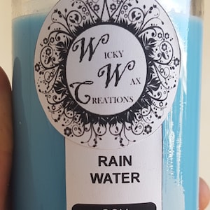 May include: A glass jar candle with a light blue wax filling. The candle has a lid and a label with the text "Rain Water Soy Candle" and the brand name "Icky Wax Creations".