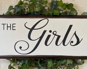 May include: A white wooden sign with a dark brown frame. The sign says "The Girls" in black cursive lettering.