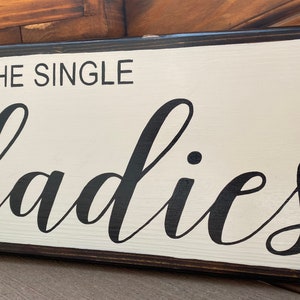 May include: A white wooden sign with black lettering that reads "All the single ladies". The sign has a dark brown wood frame.