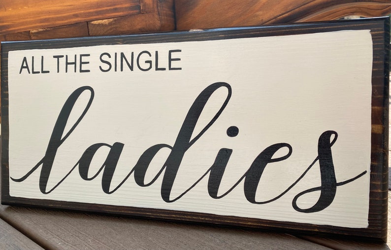May include: A white wooden sign with black lettering that reads "All the single ladies". The sign has a dark brown wood frame.