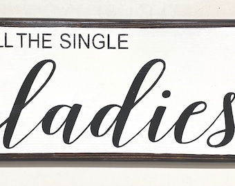 May include: A white wooden sign with a dark brown frame. The sign says "All the single ladies" in black cursive lettering.