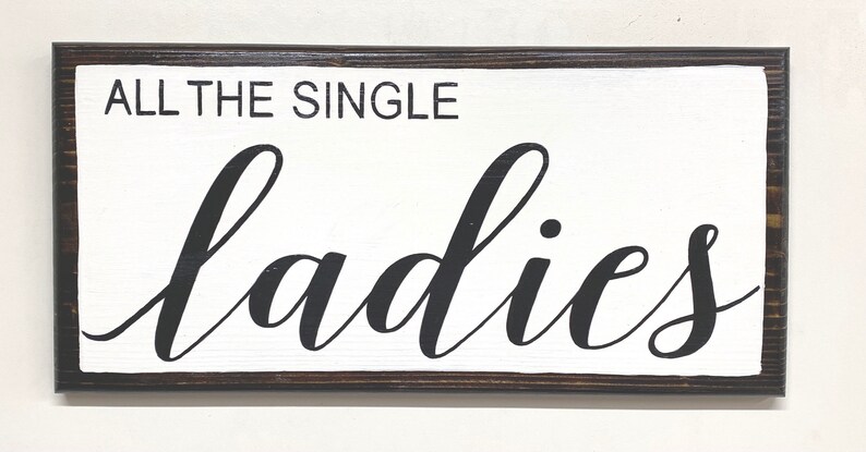 May include: A white wooden sign with a dark brown frame. The sign says "All the single ladies" in black cursive lettering.