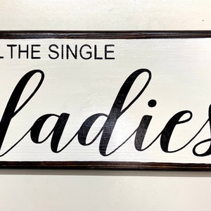 May include: A white wooden sign with black lettering that reads "All the single ladies". The sign has a dark brown frame.