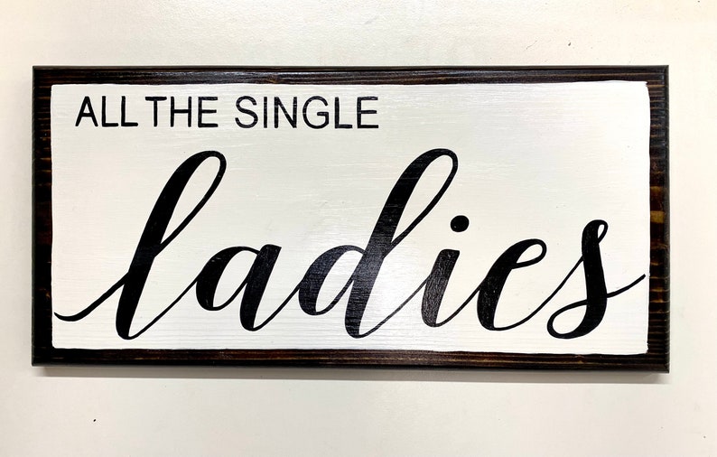 May include: A white wooden sign with black lettering that reads "All the single ladies". The sign has a dark brown frame.