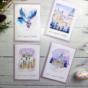May include: Four white greeting cards with watercolour illustrations of a snowy castle, a cottage, and an owl carrying a gift. Each card features the text Have a magical Christmas in black script. The cards are set against a white wooden background.
