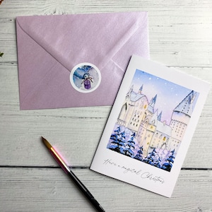 May include: A white greeting card featuring a watercolour illustration of a snow-covered castle and pine trees with the text "Have a magical Christmas," accompanied by a shimmering lavender envelope sealed with a round sticker and a black paintbrush.