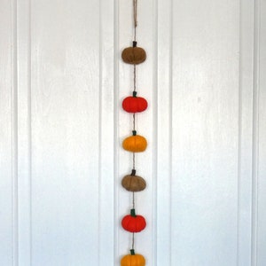 May include: A vertical hanging garland featuring six small fabric pumpkins in alternating colours of brown, orange, and yellow. Each pumpkin has a small green stem and is strung together on a thin piece of natural twine with a loop at the top.