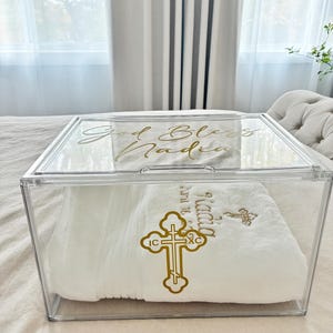 Könnte beinhalten: Eine transparente Acrylbox mit einem weißen Handtuch darin. Der Deckel der Box trägt den goldenen Schriftzug "God Bless Nadia". Das Handtuch ist mit einem goldenen Kreuz und dem Namen "Nadia" bestickt.