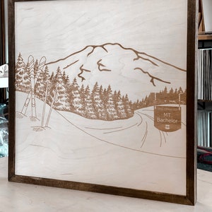 Puede incluir: Un letrero de madera con un dise&ntilde;o de corte por l&aacute;ser de una cordillera, &aacute;rboles y dos esqu&iacute;s. El letrero tiene un marco marr&oacute;n y el texto "MT. Bachelor" es visible en la esquina inferior derecha.