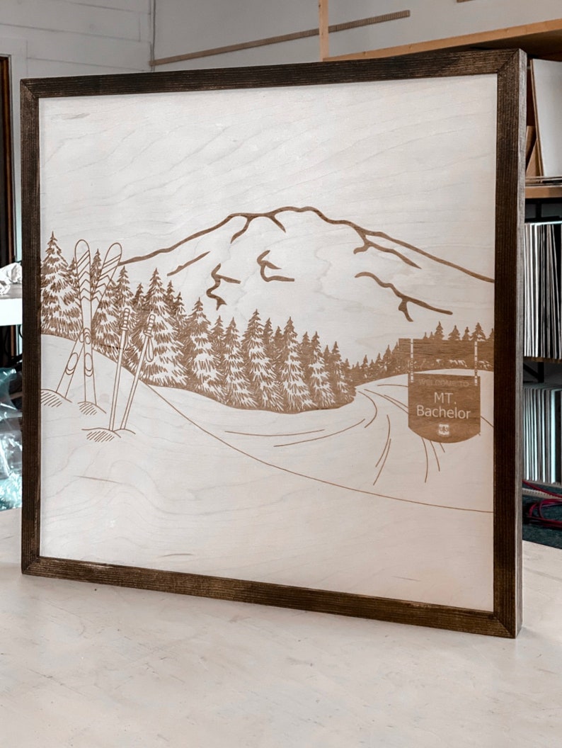 Puede incluir: Un letrero de madera con un dise&ntilde;o de corte por l&aacute;ser de una cordillera, &aacute;rboles y dos esqu&iacute;s. El letrero tiene un marco marr&oacute;n y el texto "MT. Bachelor" es visible en la esquina inferior derecha.