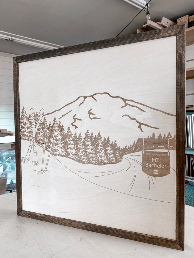 Puede incluir: Un letrero de madera con un dise&ntilde;o de monta&ntilde;a, &aacute;rboles y bastones de esqu&iacute; cortado con l&aacute;ser. El letrero dice "Bienvenido a Mt. Bachelor".
