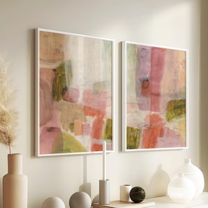 May include: Two abstract paintings with pink, green, and white colors. The paintings are framed in white and are hanging on a white wall. The paintings are above a white dresser with a vase, two round decorative objects, and a candle holder.