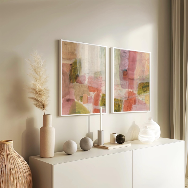 May include: Two abstract paintings with pink, green, and white colors. The paintings are framed in white and are hanging on a white wall. The paintings are above a white dresser with a vase, two round decorative objects, and a candle holder.