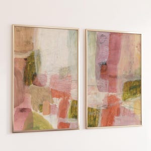 May include: Two rectangular wall art prints in thin gold frames featuring abstract compositions. The artwork displays layered washes of muted pink, soft orange, olive green, and cream tones with visible brushstrokes and thin charcoal lines.